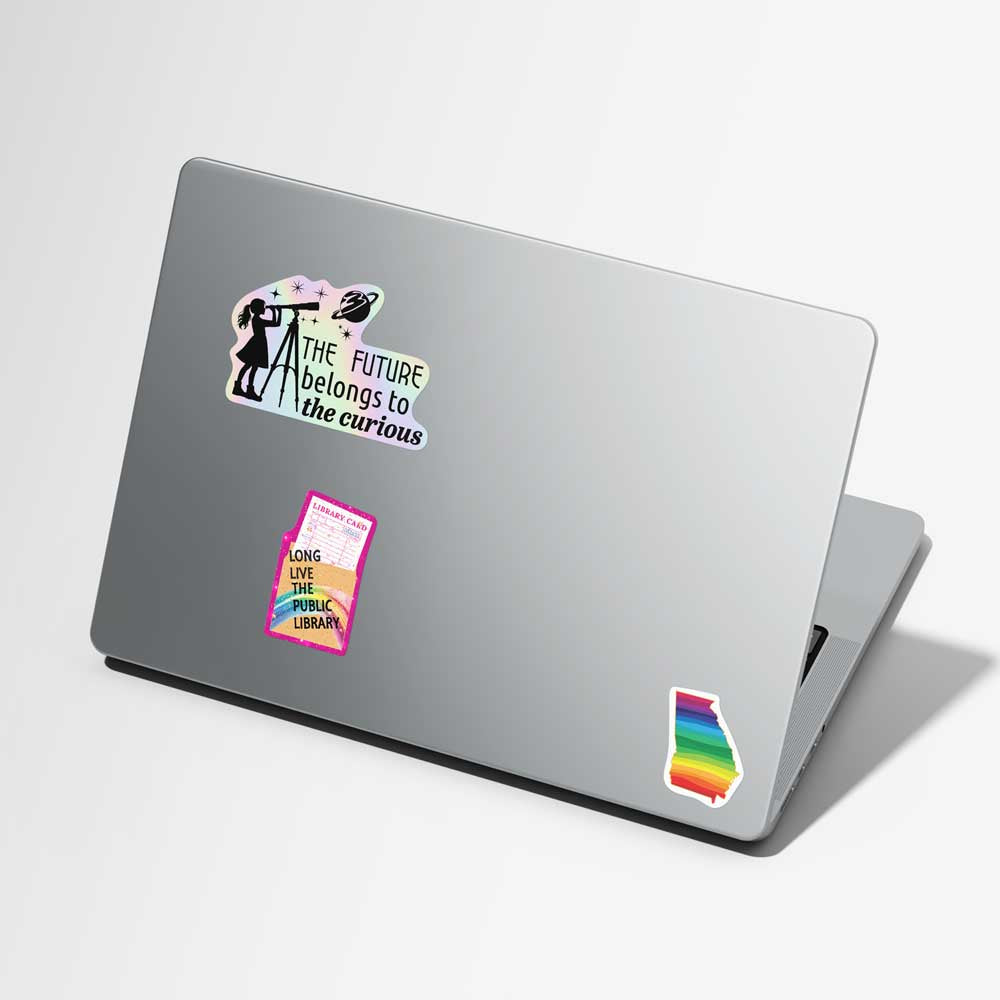 A silver laptop on a light gray background, decorated with three stickers: a black-and-white 'the future belongs to the curious' sticker, a pink and yellow 'Long Live The Librarian' book sticker, and a rainbow-striped state-of-Georgia silhouette sticker.