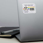 A rectangular sticker with a raccoon and rainbow design reading 'Public Library Card' and 'Everyone Welcome' is shown on the back of a silver laptop. 