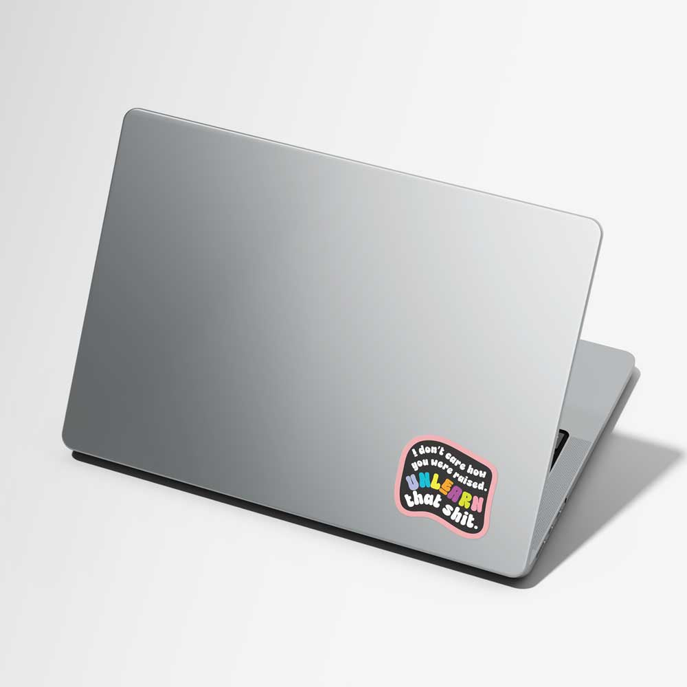 A silver laptop shown closed at an angle on a light gray background, with a colorful sticker on the lower right corner of the lid that reads 'I don't care how you were raised. Unlearn that shit'.
